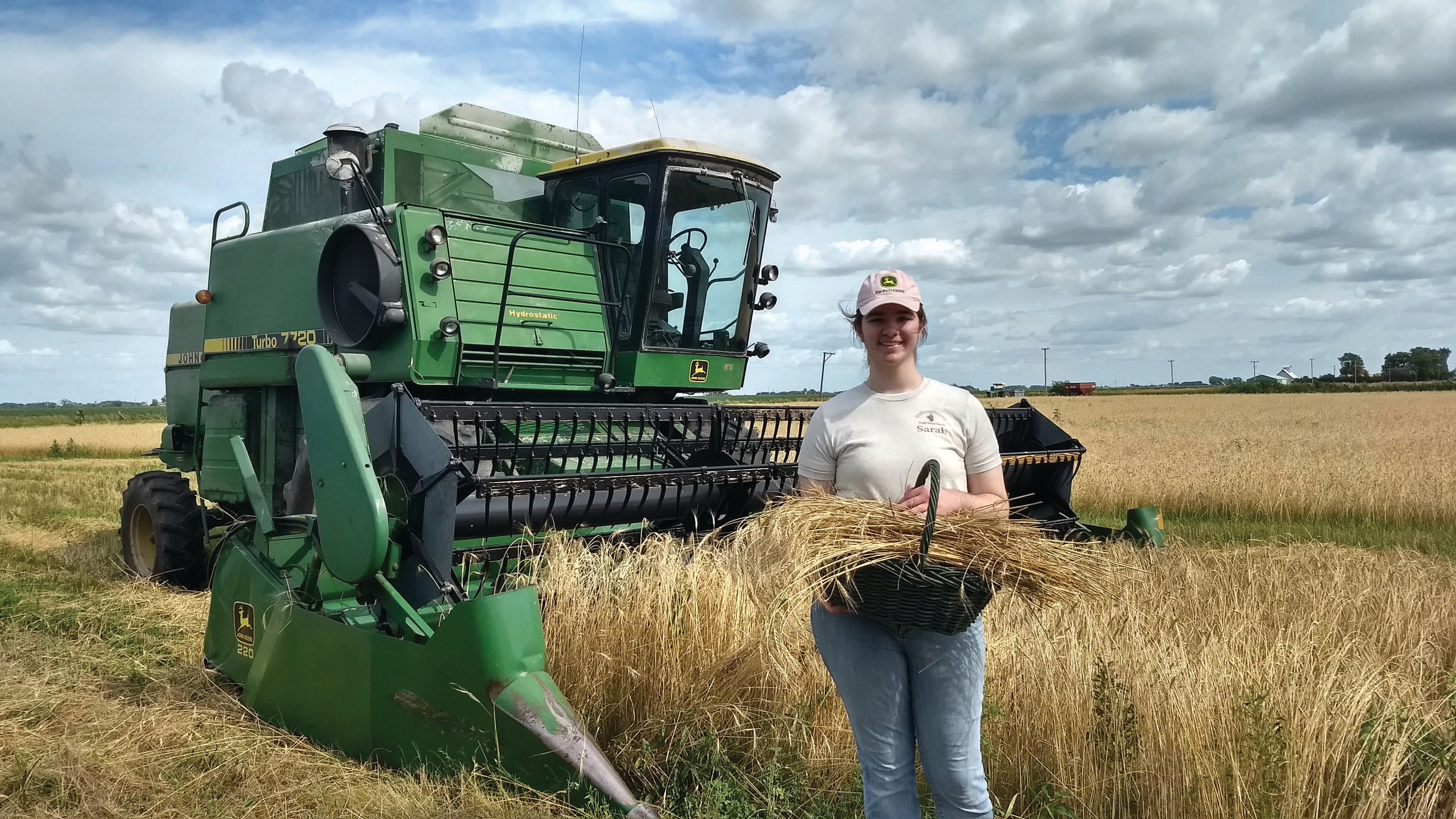 girl standing in a field with a combine harvester, holding a bundle of wheat.